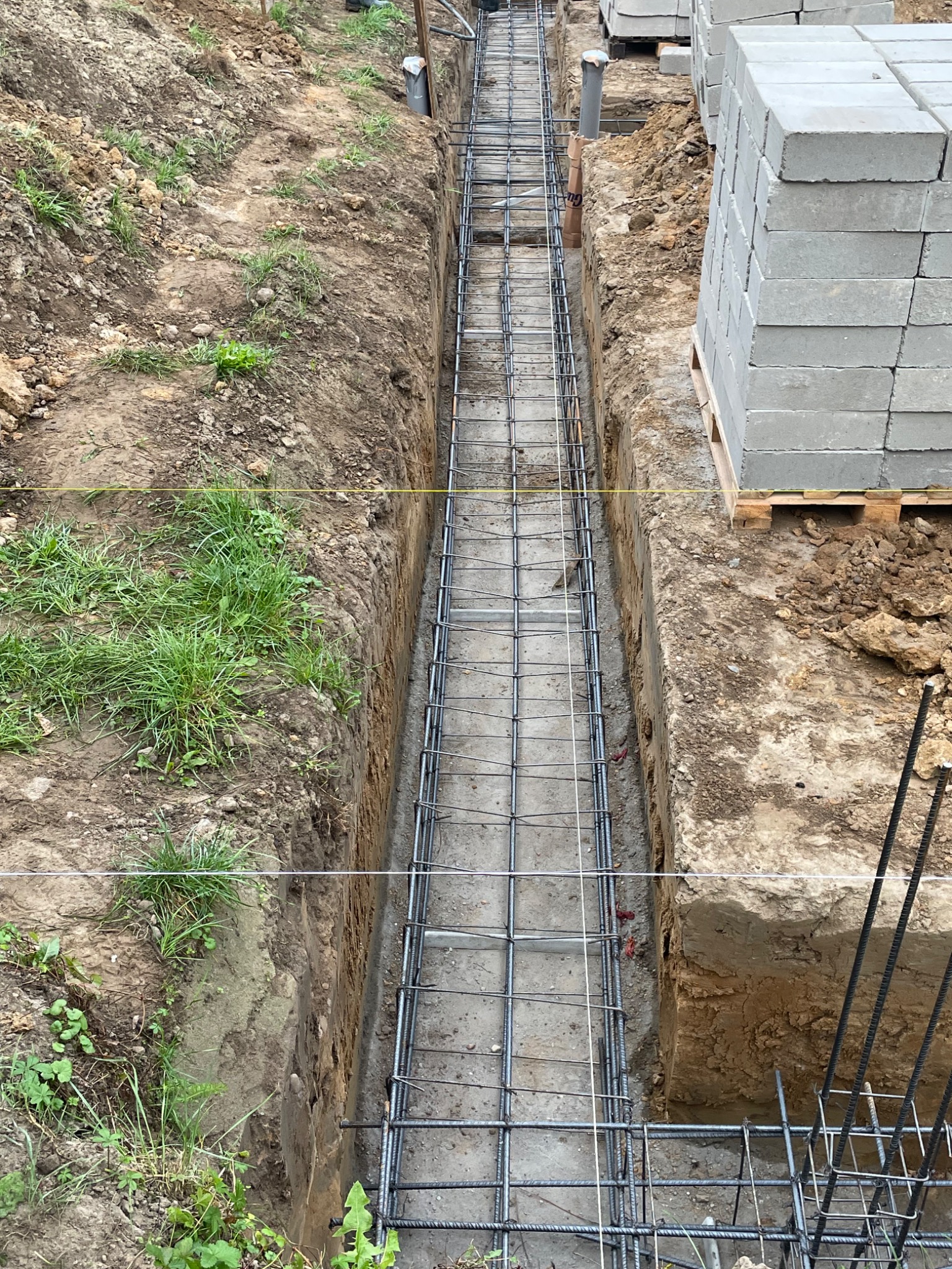 Zbrojenie w wykopie pod fundament. Widoczne ułożone pręty zbrojeniowe, sznur wyznaczający linię, paleta z bloczkami betonowymi. Grunt w wykopie.