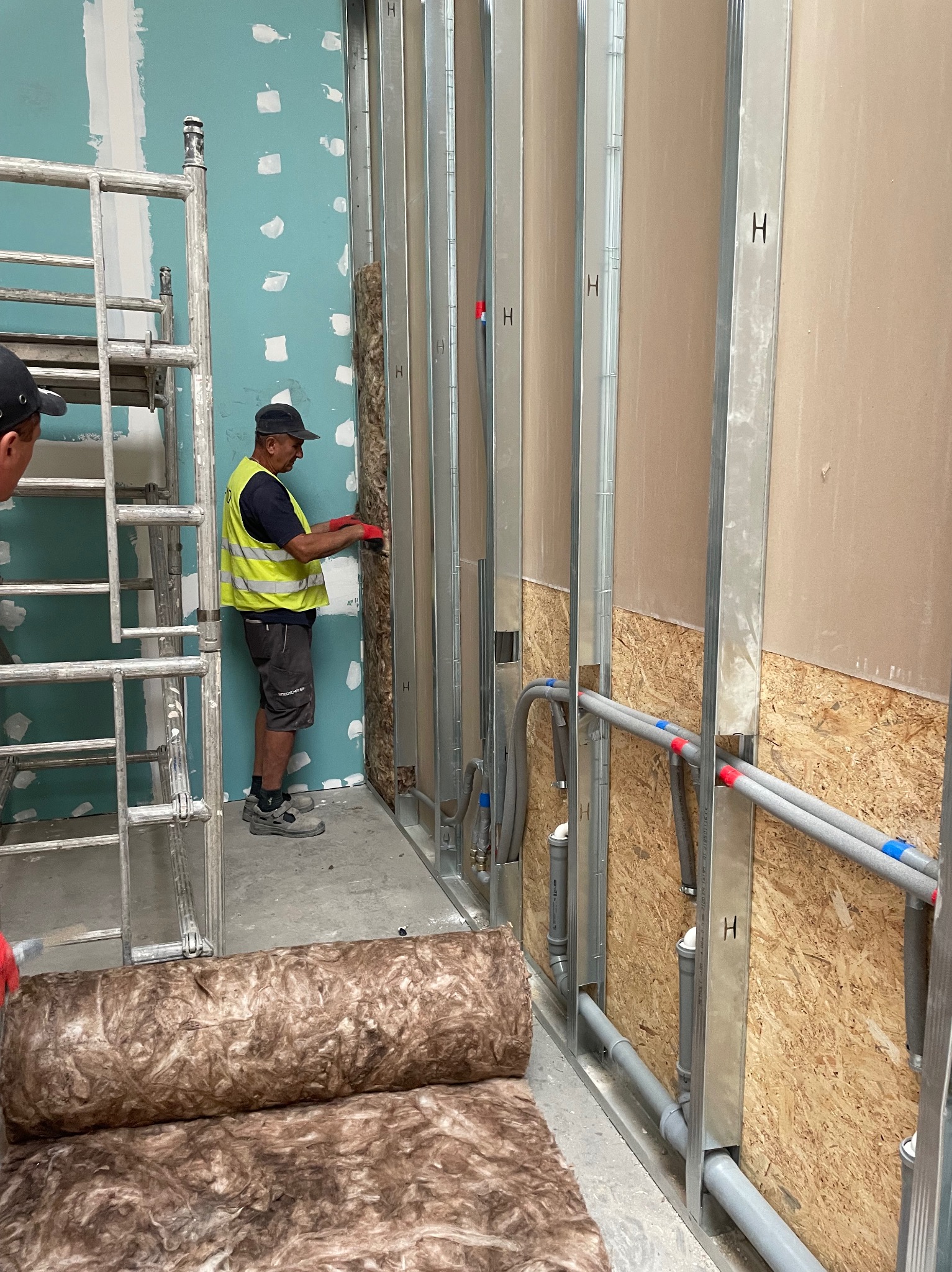 Pracownik w kamizelce montuje izolację w metalowym stelażu ściany działowej. Widoczne rolki wełny mineralnej, instalacje i fragment rusztowania.