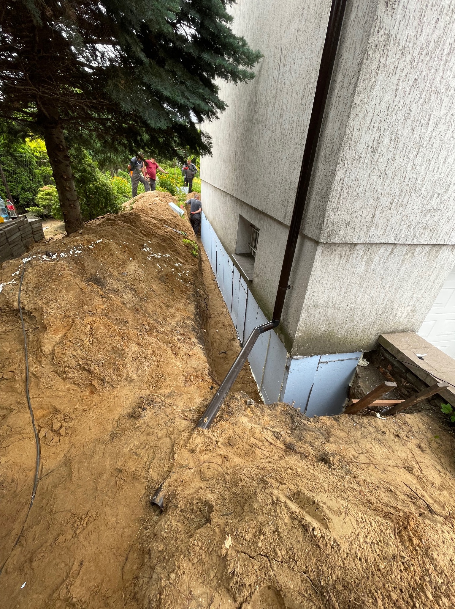 Odkopane fundamenty budynku z widoczną izolacją termiczną. W tle grupa robotników. Ujęcie z góry, ukazujące wykop i fragment elewacji.