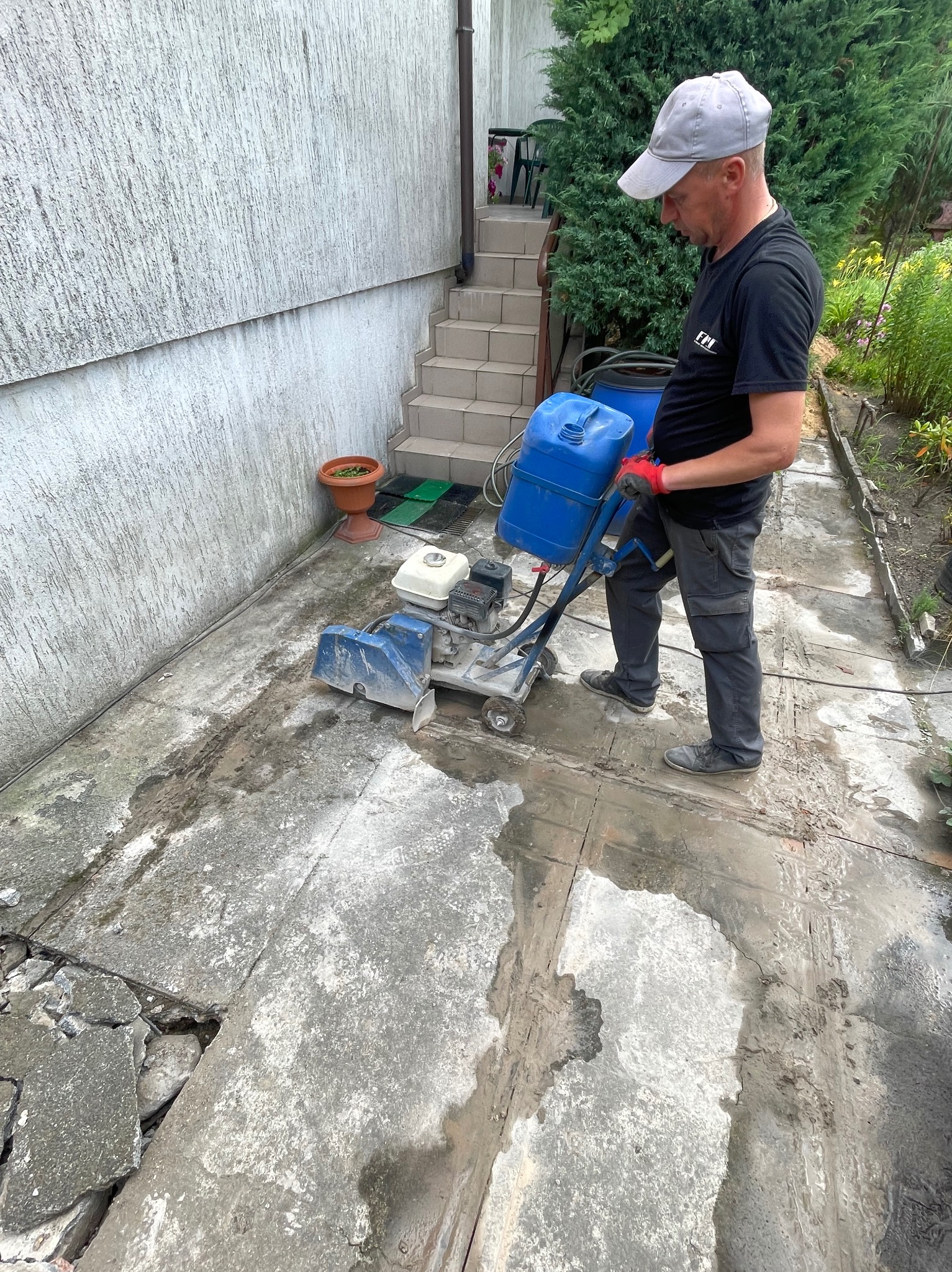 Pracownik w szarym ubraniu przecina betonową posadzkę specjalistyczną maszyną z niebieskim zbiornikiem na wodę. Widoczne pęknięcia i uszkodzenia posadzki.