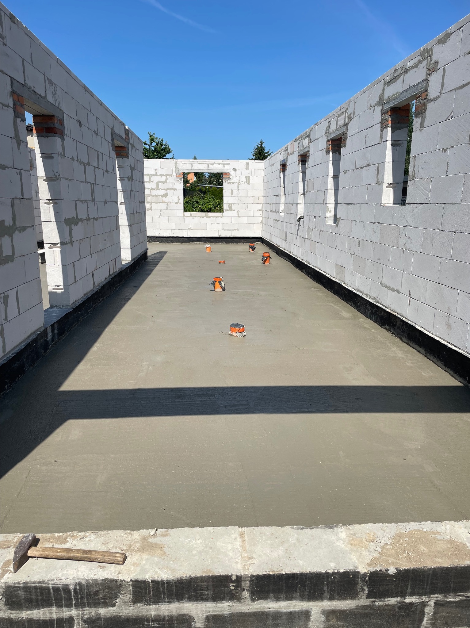 Wnętrze domu w trakcie budowy: ściany z białego bloczka, wylana posadzka z widocznymi elementami instalacji. W tle błękitne niebo i zieleń drzew.