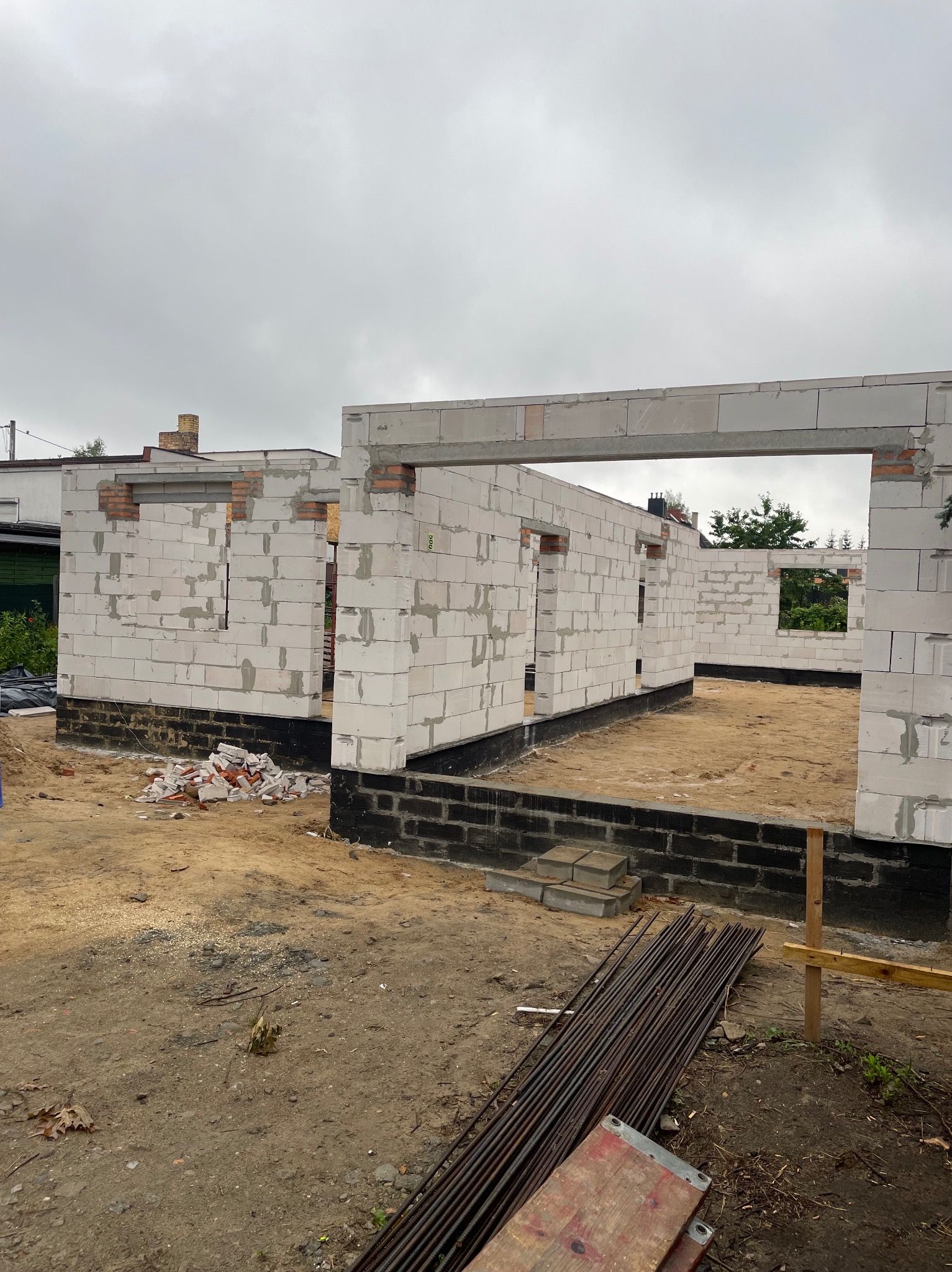 Wznoszenie ścian parteru domu z betonu komórkowego, widoczne otwory okienne i drzwiowe, czarna izolacja fundamentów, zbrojenie na ziemi. Pochmurne niebo w tle.