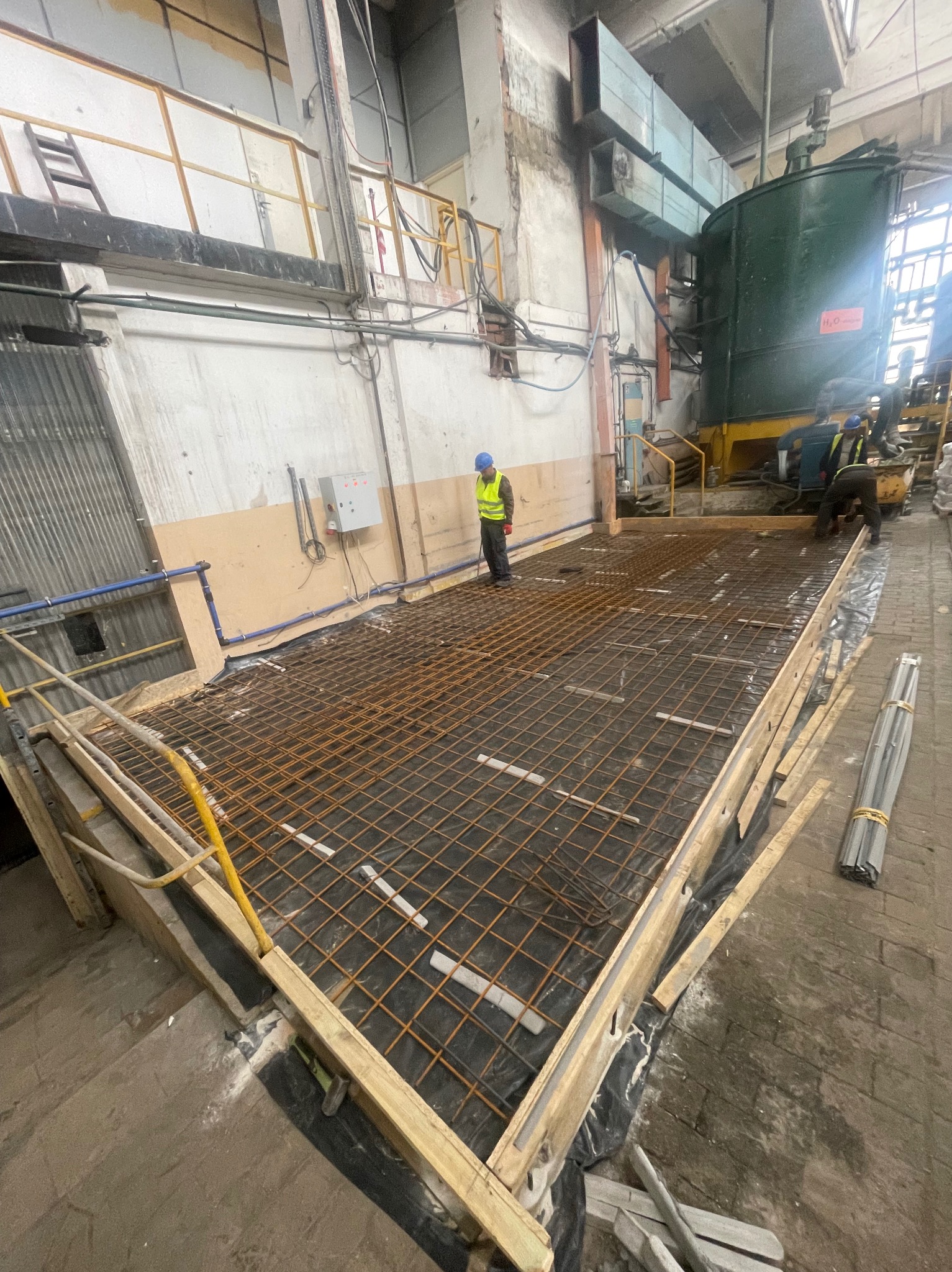 Zbrojenie podłoża w hali przemysłowej, widoczne siatki zbrojeniowe, szalunki drewniane i pracownicy w kaskach. Przygotowanie do wylewki betonowej.