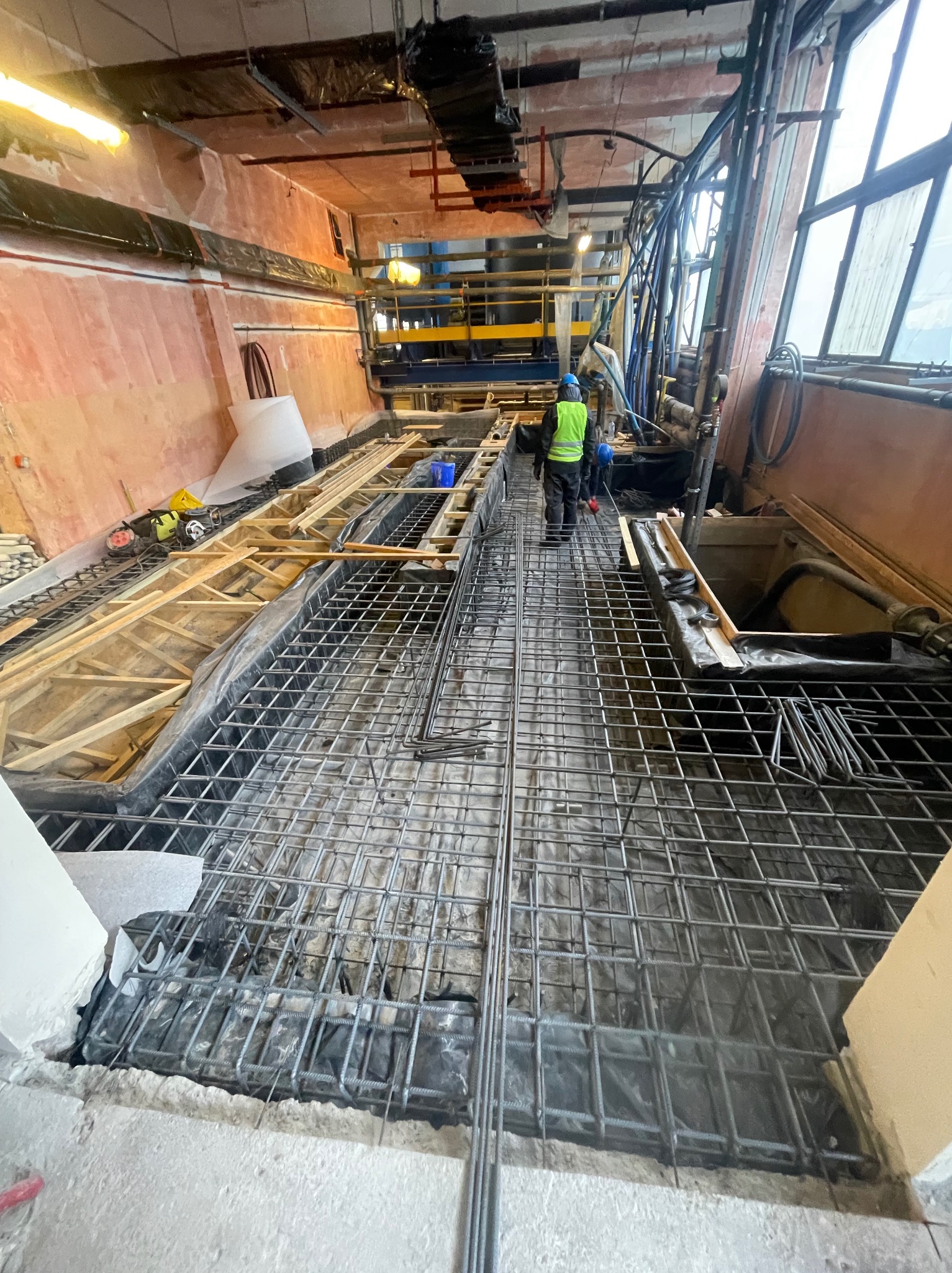Zbrojenie fundamentów z siatki stalowej na folii, widoczny pracownik w kamizelce odblaskowej i niebieskim kasku, drewniane szalunki w tle, surowy stan budynku.