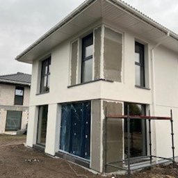 MirkoPol Mirosław Snarski - Biały dom w trakcie ocieplania: szare płyty izolacyjne wokół okien, częściowo otynkowana elewacja, widoczne rusztowanie. Dom z dwuspadowym dachem, sąsiedni budynek w stanie surowym.