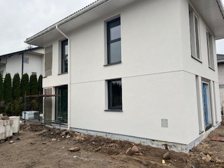 Biały, dwukondygnacyjny dom z szarymi oknami w trakcie wykończenia elewacji. Widoczne rusztowanie i materiały budowlane. Surowy teren wokół budynku.