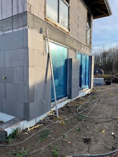 Ocieplanie ścian zewnętrznych budynku z szarego styropianu, widoczne okna zabezpieczone folią, przewody elektryczne na ziemi, w tle zieleń.