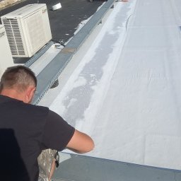 HydroRoof Hydroizolacje Dachów Płaskich Płynnymi Membranami Patryk Przekwas - Pracownik w czarnej koszulce HydroRoof uszczelnia białą membranę dachu płaskiego. Widoczne klimatyzatory i metalowe elementy konstrukcyjne na dachu.