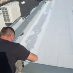 Pracownik w czarnej koszulce HydroRoof uszczelnia białą membranę dachu płaskiego. Widoczne klimatyzatory i metalowe elementy konstrukcyjne na dachu.
