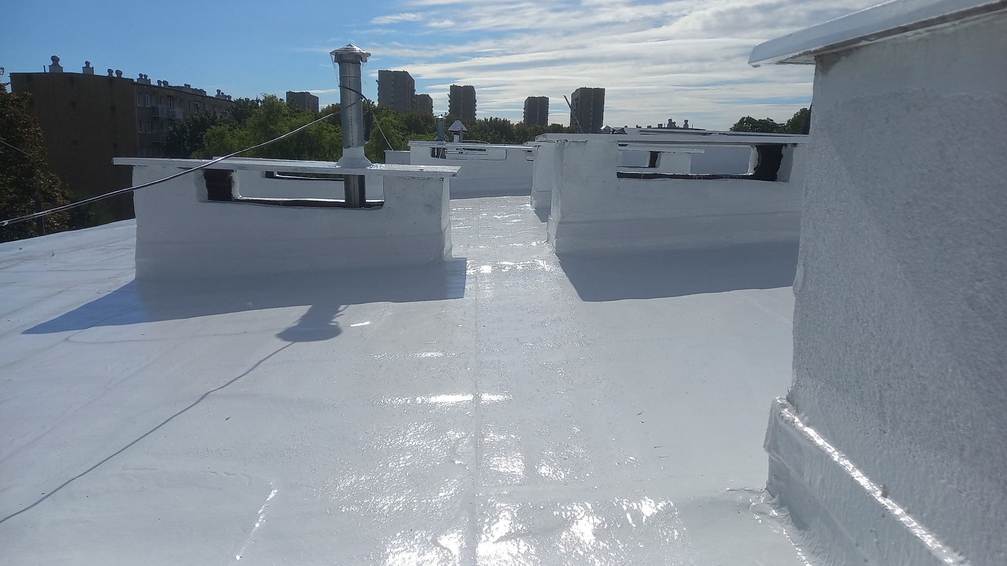 Biała, płaska powierzchnia dachu po renowacji z widocznymi kominami wentylacyjnymi i budynkami w tle, w słoneczny dzień. Powłoka wydaje się być świeżo nałożona.