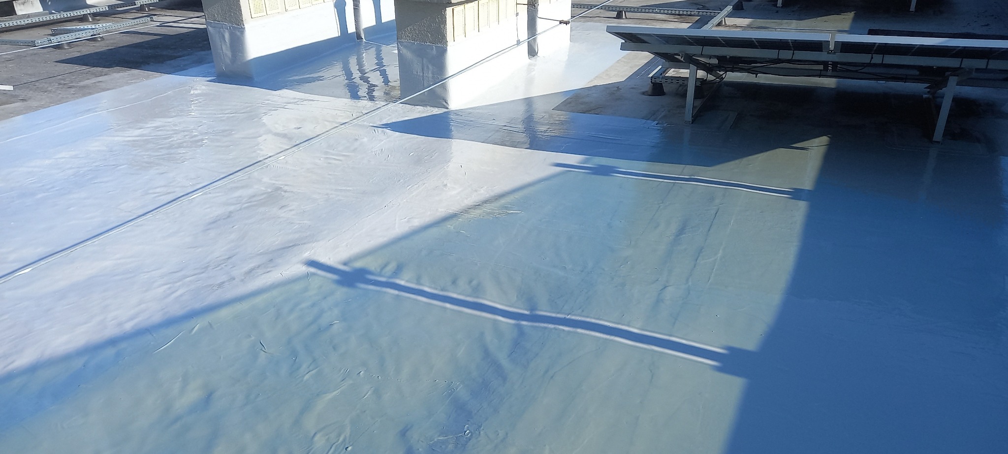 Świeżo nałożona, połyskliwa hydroizolacja w kolorze szaro-niebieskim na płaskim dachu z widocznymi elementami konstrukcyjnymi i panelami słonecznymi w tle.