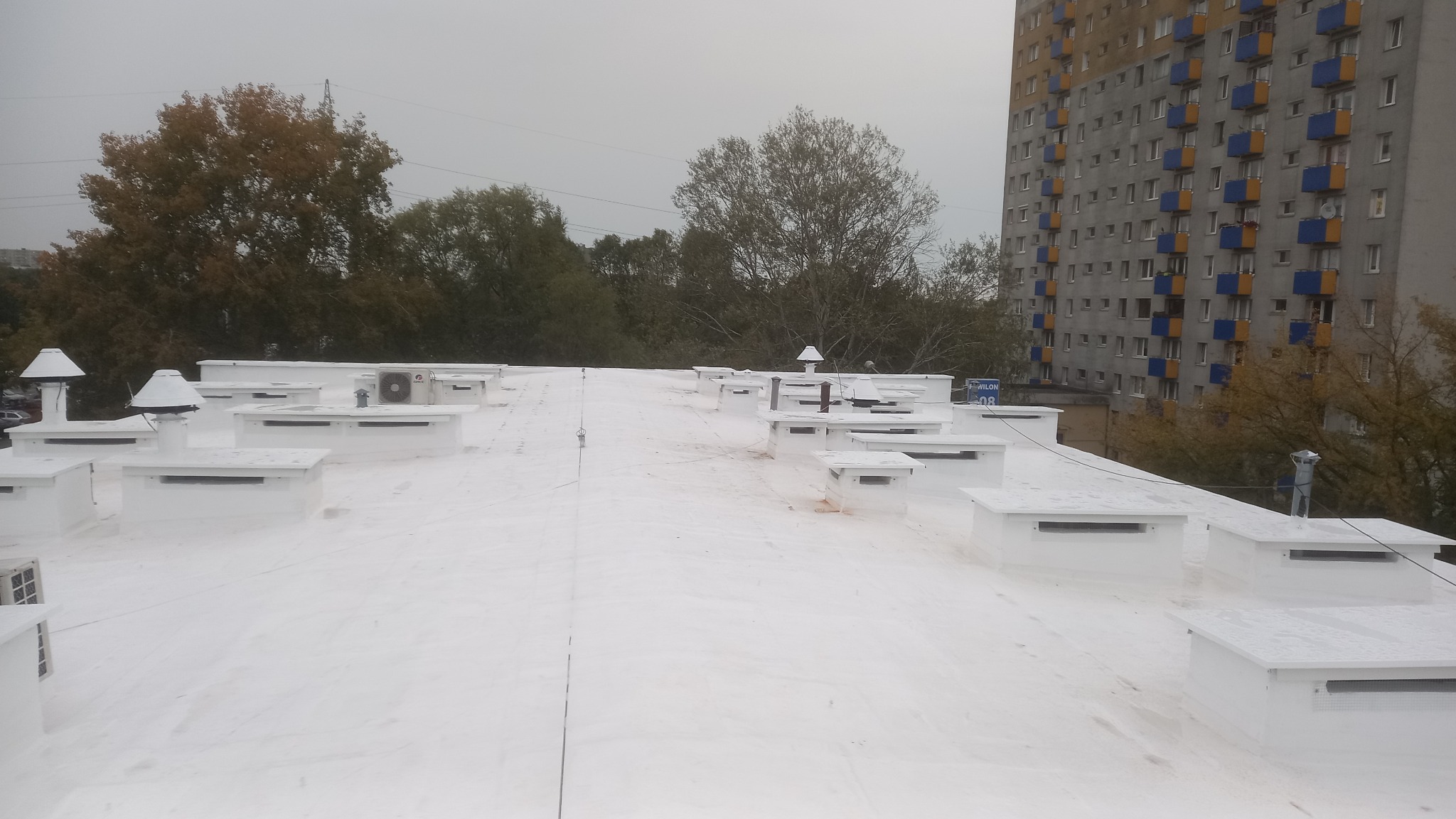 Biały dach płaski z licznymi wywietrznikami i klimatyzatorami, widok z góry na tle drzew i bloku mieszkalnego w pochmurny dzień. Widoczny adres na dachu.