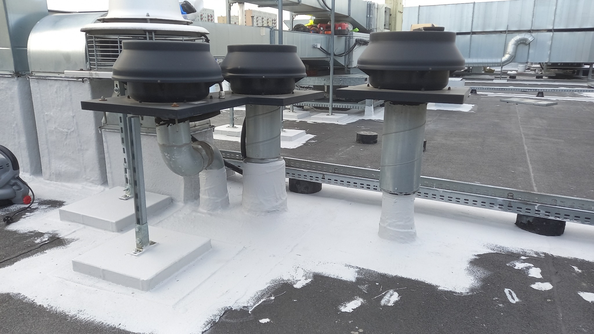 Wykończony dach płaski z trzema kominami wentylacyjnymi, zabezpieczonymi białą powłoką hydroizolacyjną. Widoczne elementy instalacji i konstrukcji wsporcze.