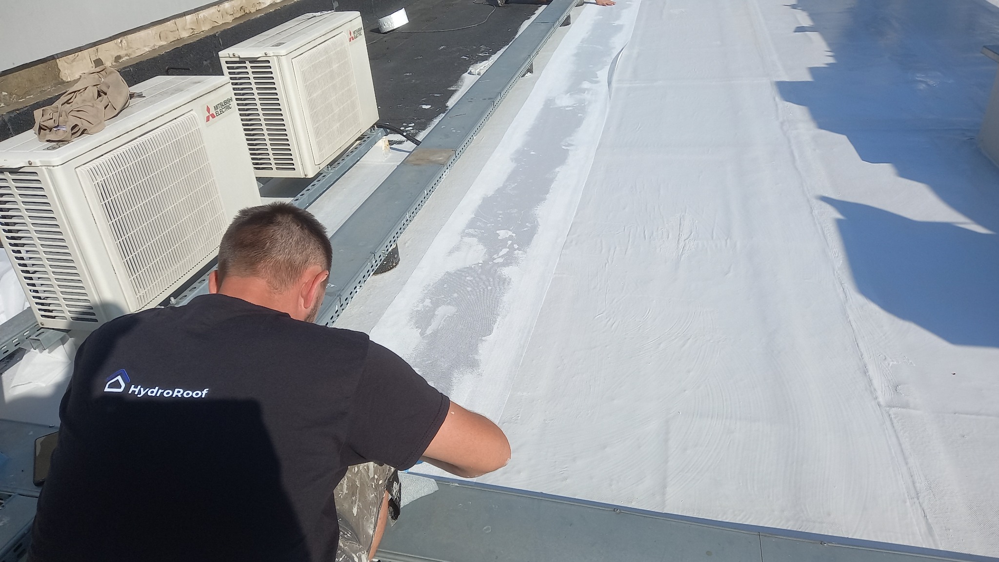 Pracownik w czarnej koszulce HydroRoof uszczelnia białą membranę dachu płaskiego. Widoczne klimatyzatory i metalowe elementy konstrukcyjne na dachu.