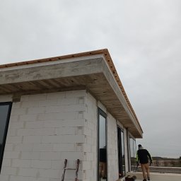 URBAN ALPSERWIS SPÓŁKA Z OGRANICZONĄ ODPOWIEDZIALNOŚCIĄ - Budynek w trakcie budowy, ściany z białych bloczków, zamontowane okna, betonowy okap dachu z drewnianym wykończeniem, drabina oparta o ścianę, pracownik na dachu w oddali.