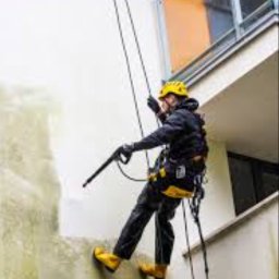 URBAN ALPSERWIS SPÓŁKA Z OGRANICZONĄ ODPOWIEDZIALNOŚCIĄ - Pracownik zabezpieczony linami, w kasku i żółtych butach, czyści elewację budynku myjką ciśnieniową, widoczny efekt czyszczenia.