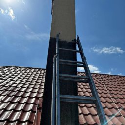 URBAN ALPSERWIS SPÓŁKA Z OGRANICZONĄ ODPOWIEDZIALNOŚCIĄ - Metalowa drabina oparta o komin na tle czerwonego dachu i błękitnego nieba. Część komina pomalowana na ciemno, kontrastująca z resztą w jasnym kolorze.