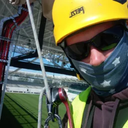 Pracownik w kasku i okularach, z zabezpieczeniem linowym na tle konstrukcji stadionu i zielonej murawy. Ujęcie z perspektywy osoby wykonującej prace na wysokości.