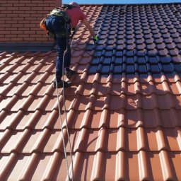 URBAN ALPSERWIS SPÓŁKA Z OGRANICZONĄ ODPOWIEDZIALNOŚCIĄ - Pracownik w uprzęży maluje dachówkę na ciemny kolor. Widoczny fragment dachu przed i po malowaniu. Dachówka ceramiczna. Ceglany komin w tle.