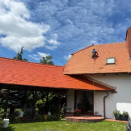 Pracownik na dachu domu z czerwoną dachówką, zabezpieczony liną. Widoczne narzędzia na trawie, dachówka i fragment elewacji. Błękitne niebo z chmurami w tle.