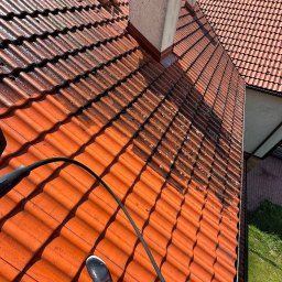 URBAN ALPSERWIS SPÓŁKA Z OGRANICZONĄ ODPOWIEDZIALNOŚCIĄ - Częściowo oczyszczony dach z czerwonej dachówki ceramicznej, widoczny fragment węża ciśnieniowego i but pracownika. Dach z kominem i widocznym fragmentem budynku.
