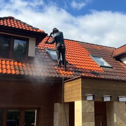 URBAN ALPSERWIS SPÓŁKA Z OGRANICZONĄ ODPOWIEDZIALNOŚCIĄ - Mężczyzna w uprzęży myje dach z czerwonej dachówki za pomocą myjki ciśnieniowej. Widoczna różnica czystości dachu. Błękitne niebo z chmurami w tle.