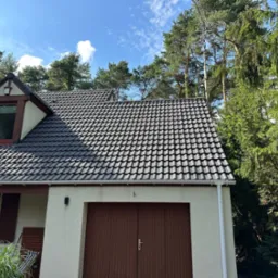 Ciemna dachówka ceramiczna na jasnym domu z brązową bramą garażową, otoczonym zielenią drzew i błękitnym niebem z chmurami. Widok z dołu.