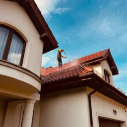 Pracownik w uprzęży czyści czerwoną dachówkę domu jednorodzinnego za pomocą myjki ciśnieniowej. Widok od dołu na tle błękitnego nieba.