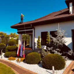 Pracownik w uprzęży na brązowym dachu domu podczas prac. Widoczne flagi i ozdobne krzewy w otoczeniu budynku.