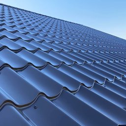 URBAN ALPSERWIS SPÓŁKA Z OGRANICZONĄ ODPOWIEDZIALNOŚCIĄ - Błyszczący, ciemny dach z blachy trapezowej pod błękitnym niebem, widok z dołu. Widoczne detale łączeń i fragment drzewa w tle.