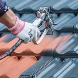 URBAN ALPSERWIS SPÓŁKA Z OGRANICZONĄ ODPOWIEDZIALNOŚCIĄ - Malowanie dachówki pistoletem natryskowym. Widoczna pomarańczowa i szara dachówka. Ręka w rękawicy trzyma pistolet. Detal z bliska, ostre światło.