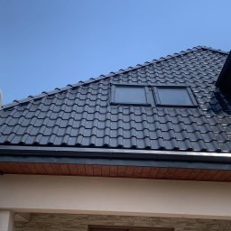 URBAN ALPSERWIS SPÓŁKA Z OGRANICZONĄ ODPOWIEDZIALNOŚCIĄ - Błyszczący, czarny dach z dachówką ceramiczną i dwoma oknami dachowymi, widziany z dołu pod kątem. Na pierwszym planie antena satelitarna i fragment elewacji budynku.