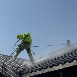 URBAN ALPSERWIS SPÓŁKA Z OGRANICZONĄ ODPOWIEDZIALNOŚCIĄ - Pracownik w jaskrawozielonym kombinezonie, zabezpieczony linami, czyści dach z ciemnej dachówki za pomocą myjki ciśnieniowej. Woda spływa po dachu.