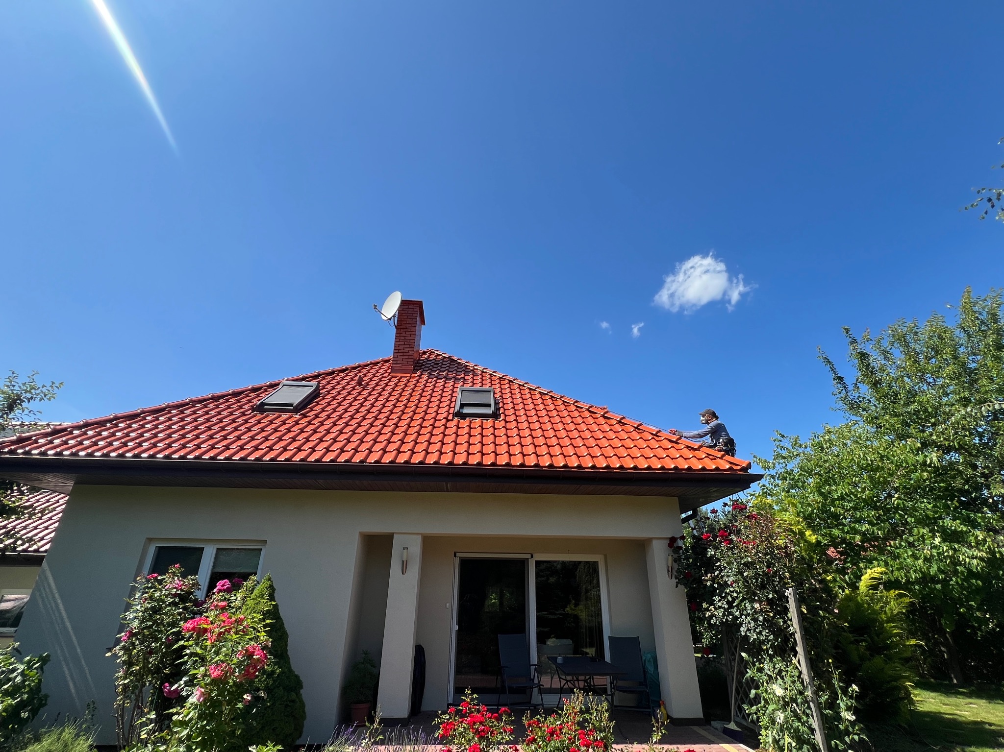 Czerwony dach z dachówki ceramicznej, na którym pracuje dekarz w szarej odzieży, widoczny na tle błękitnego nieba i zielonej roślinności.