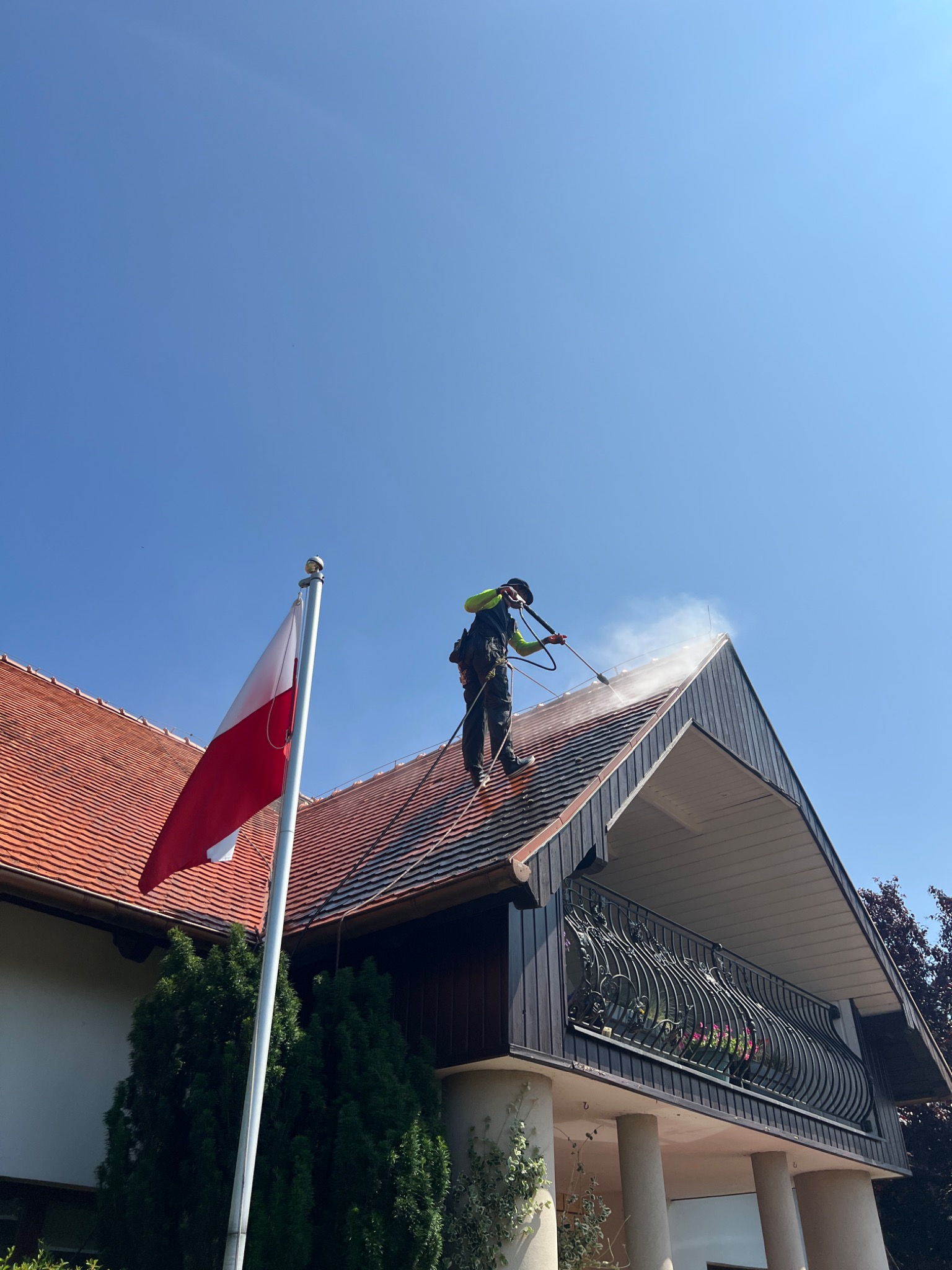 Pracownik w uprzęży czyści dach domu z flagą Polski, używając myjki ciśnieniowej. Widok z dołu na budynek z balkonem i czerwoną dachówką.