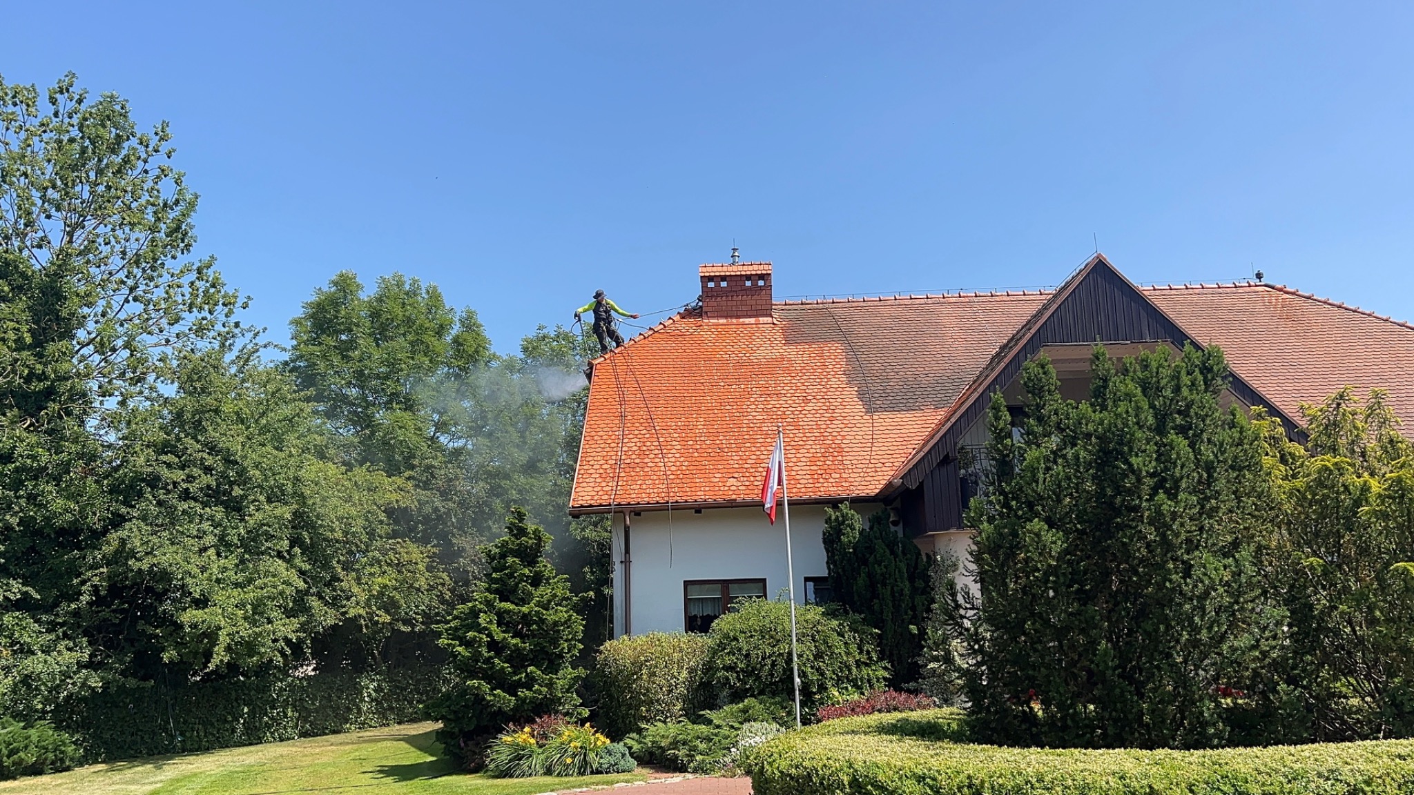 Pracownik w uprzęży czyści dach z czerwonej dachówki myjką ciśnieniową. Widoczny efekt czyszczenia dachu. W tle drzewa i budynek z polską flagą.