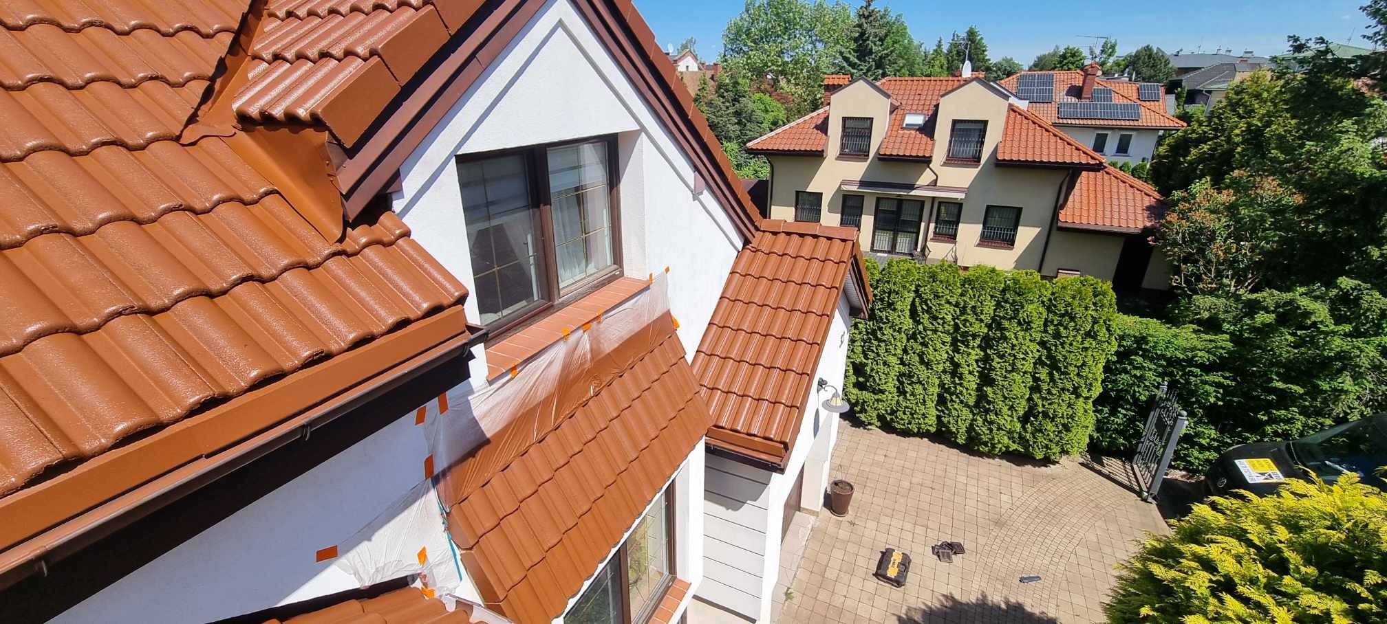 Błyszczący, brązowy dach po malowaniu, z zabezpieczonymi oknami folią. Widok z góry na dom z ogrodem i kostką brukową. Słoneczny dzień.