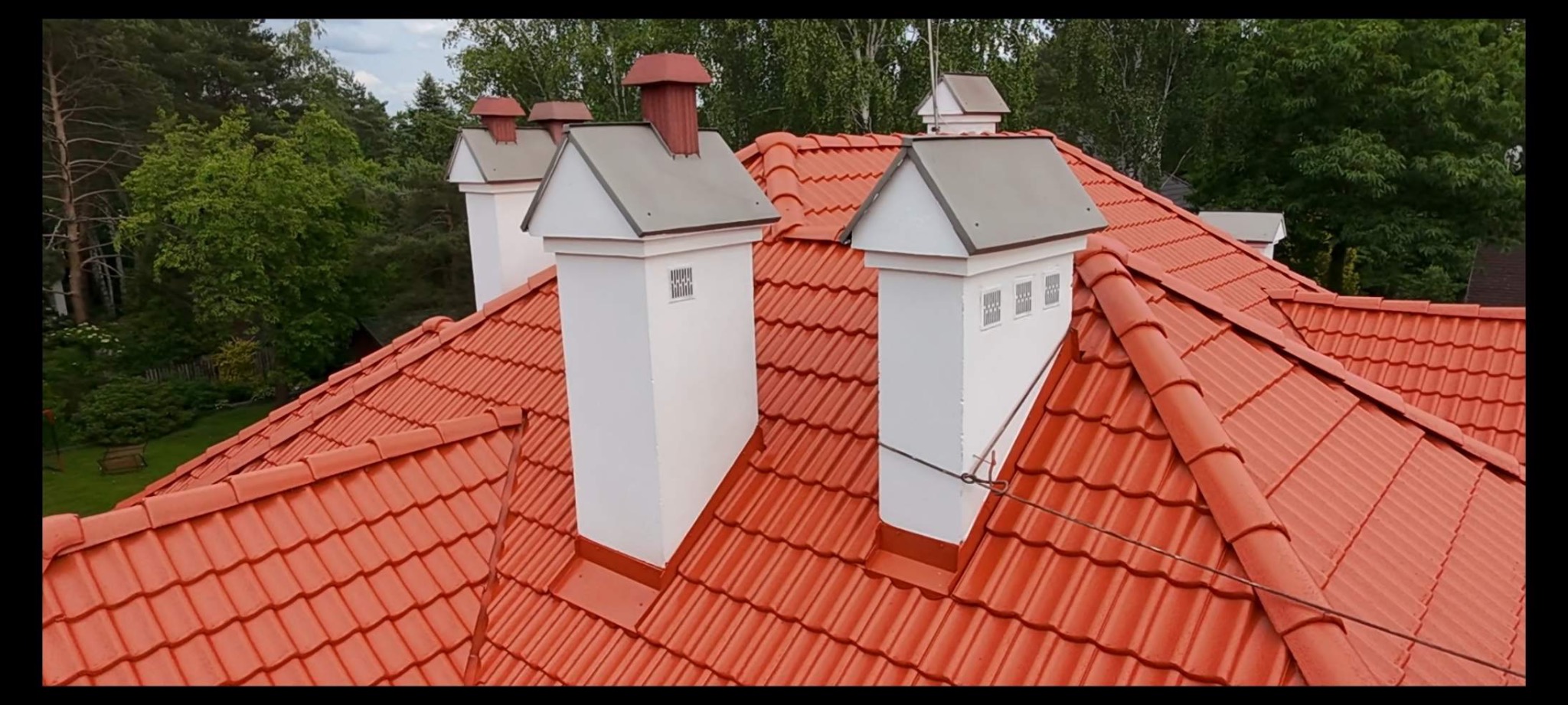 Czerwony dach z dachówki ceramicznej z białymi kominami na tle zielonych drzew. Widok z góry na odnowiony dach.