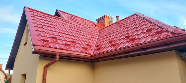 Czerwony dach z blachodachówki z nowymi rynnami w odcieniu bordo na tle błękitnego nieba i fragmentu elewacji domu w kolorze beżowym.