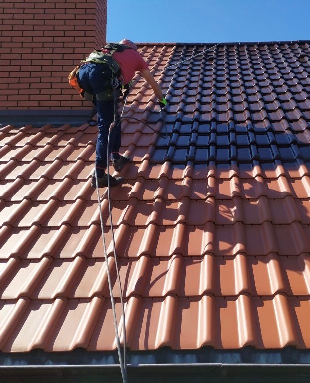 Pracownik w uprzęży maluje dachówkę na ciemny kolor. Widoczny fragment dachu przed i po malowaniu. Dachówka ceramiczna. Ceglany komin w tle.