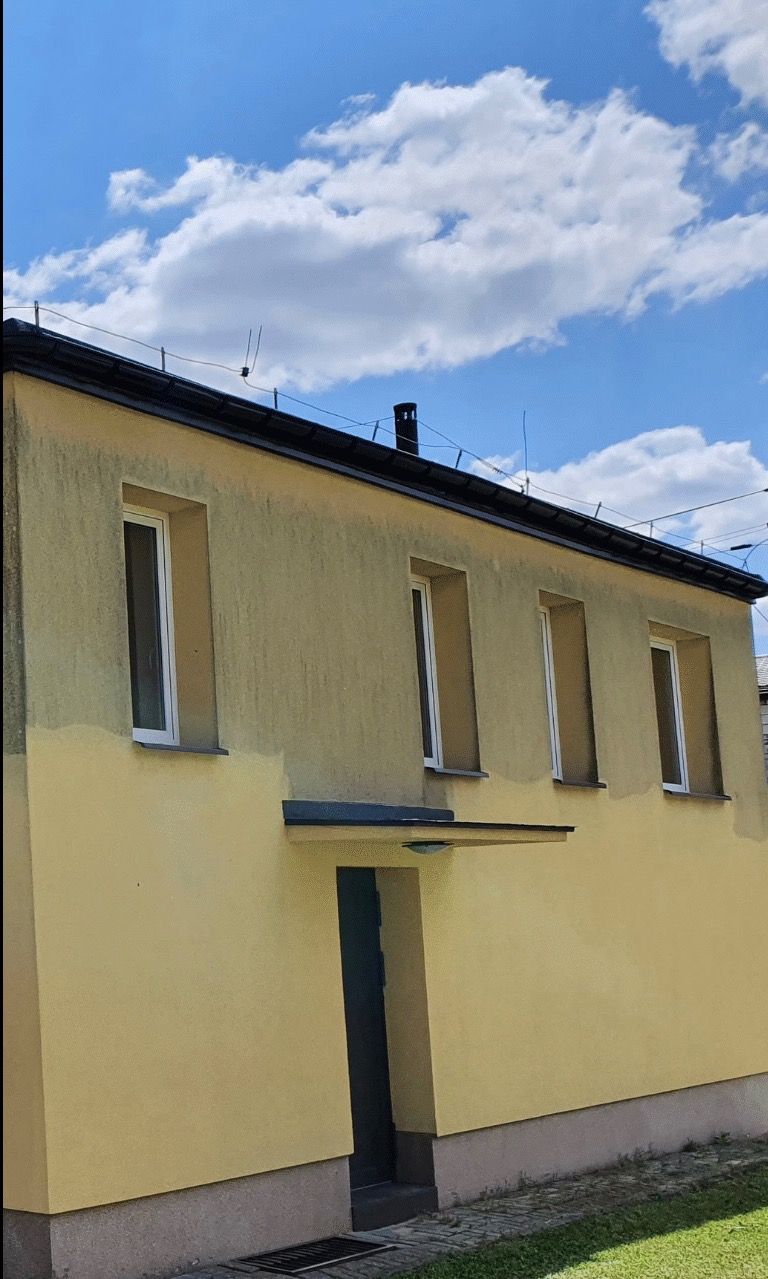 Żółta elewacja budynku w trakcie renowacji, widoczne fragmenty świeżo pomalowanej ściany, okna z białymi ramami, na dachu antena.