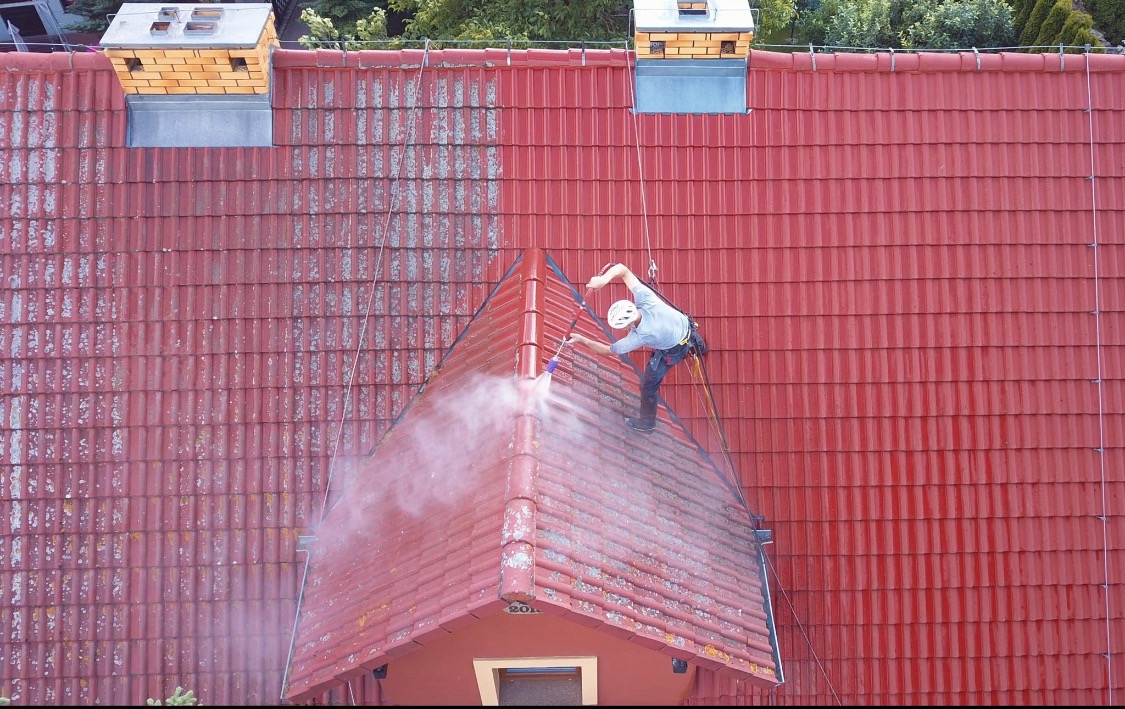Pracownik w uprzęży czyści czerwoną dachówkę myjką ciśnieniową. Widoczny kontrast między czystą a zabrudzoną powierzchnią dachu. Ujęcie z góry.
