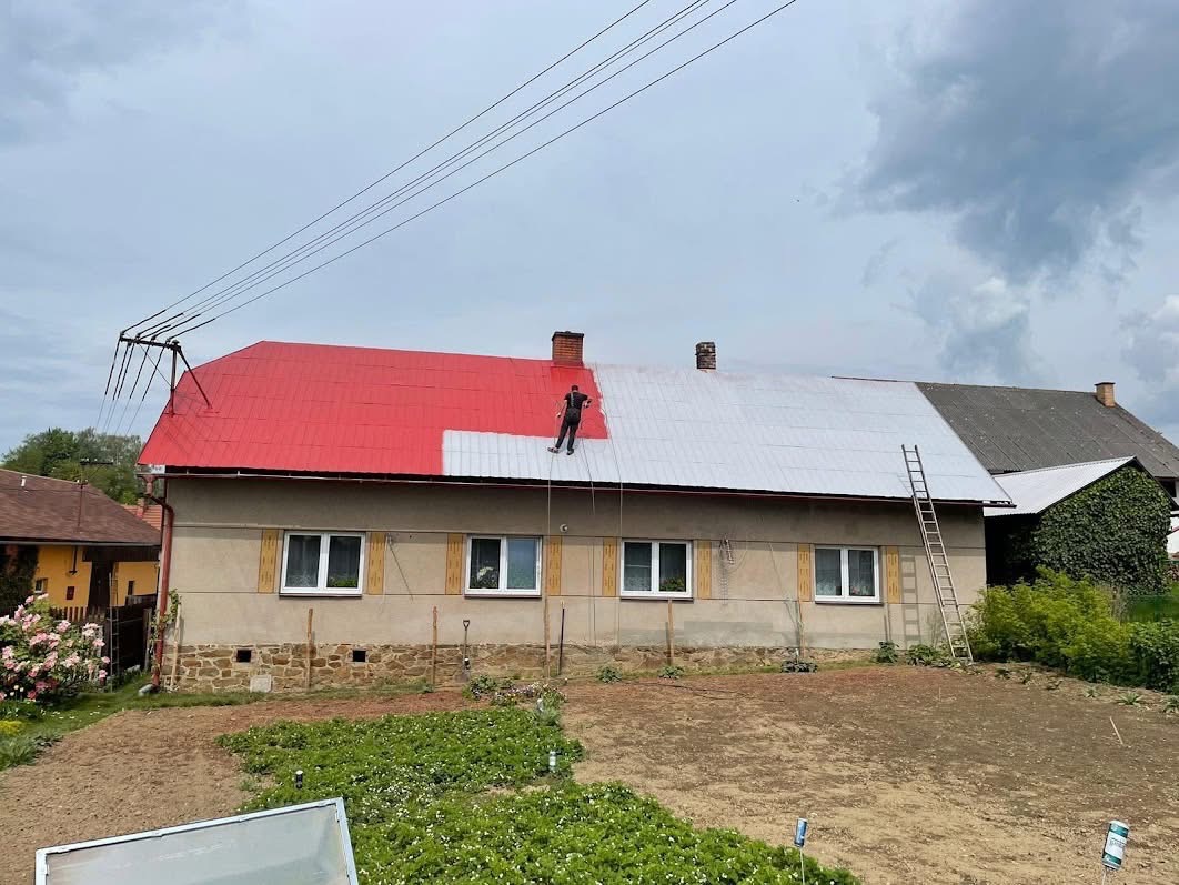 Malowanie dachu domu jednorodzinnego na czerwono i szaro. Prace na wysokości z użyciem liny. Częściowo pomalowany dach. Drabina oparta o budynek.