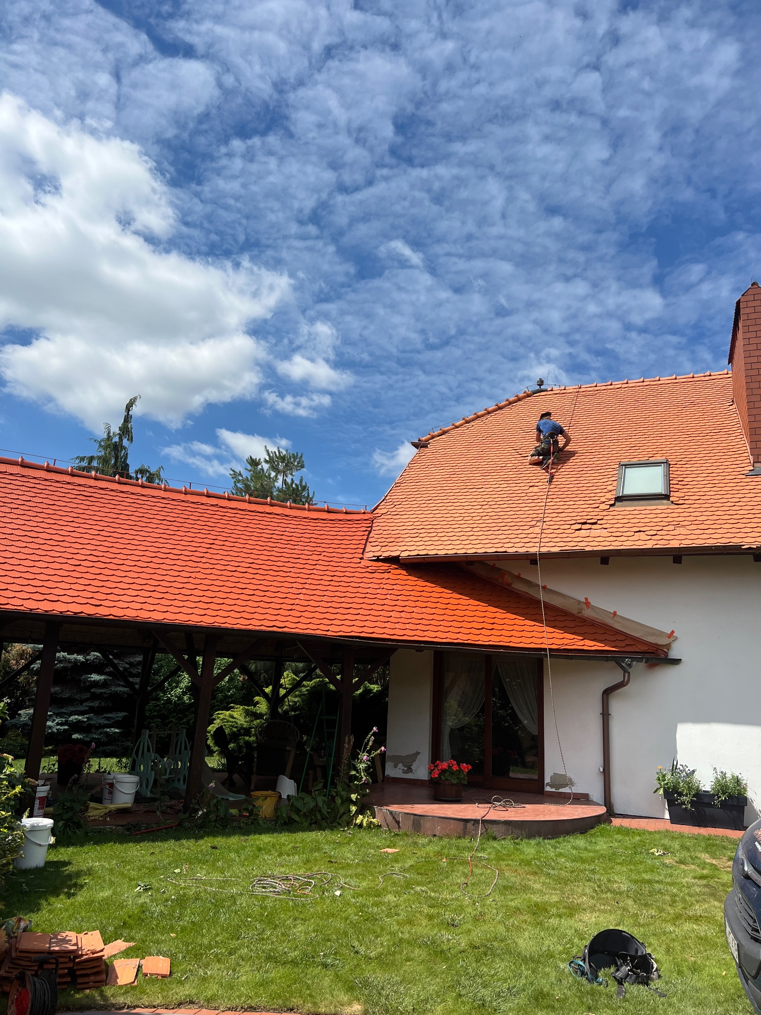 Pracownik na dachu domu z czerwoną dachówką, zabezpieczony liną. Widoczne narzędzia na trawie, dachówka i fragment elewacji. Błękitne niebo z chmurami w tle.