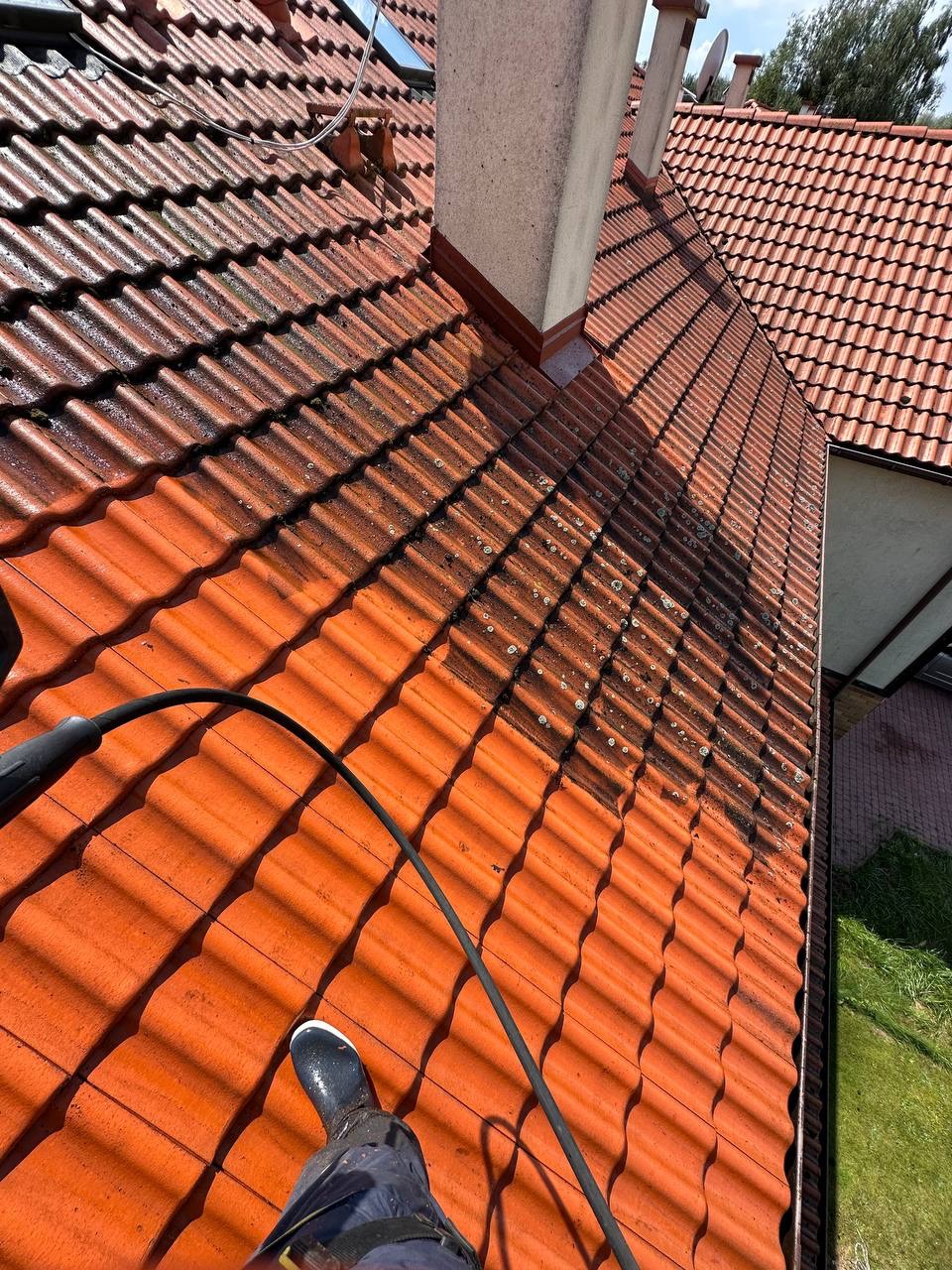 Częściowo oczyszczony dach z czerwonej dachówki ceramicznej, widoczny fragment węża ciśnieniowego i but pracownika. Dach z kominem i widocznym fragmentem budynku.