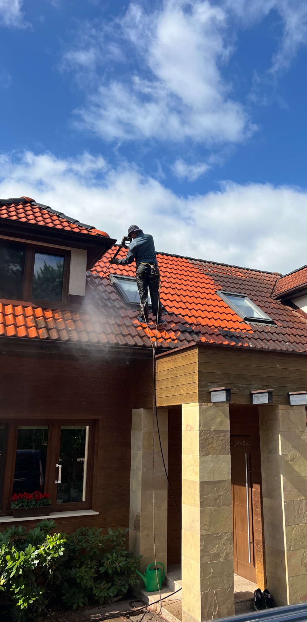 Mężczyzna w uprzęży myje dach z czerwonej dachówki za pomocą myjki ciśnieniowej. Widoczna różnica czystości dachu. Błękitne niebo z chmurami w tle.