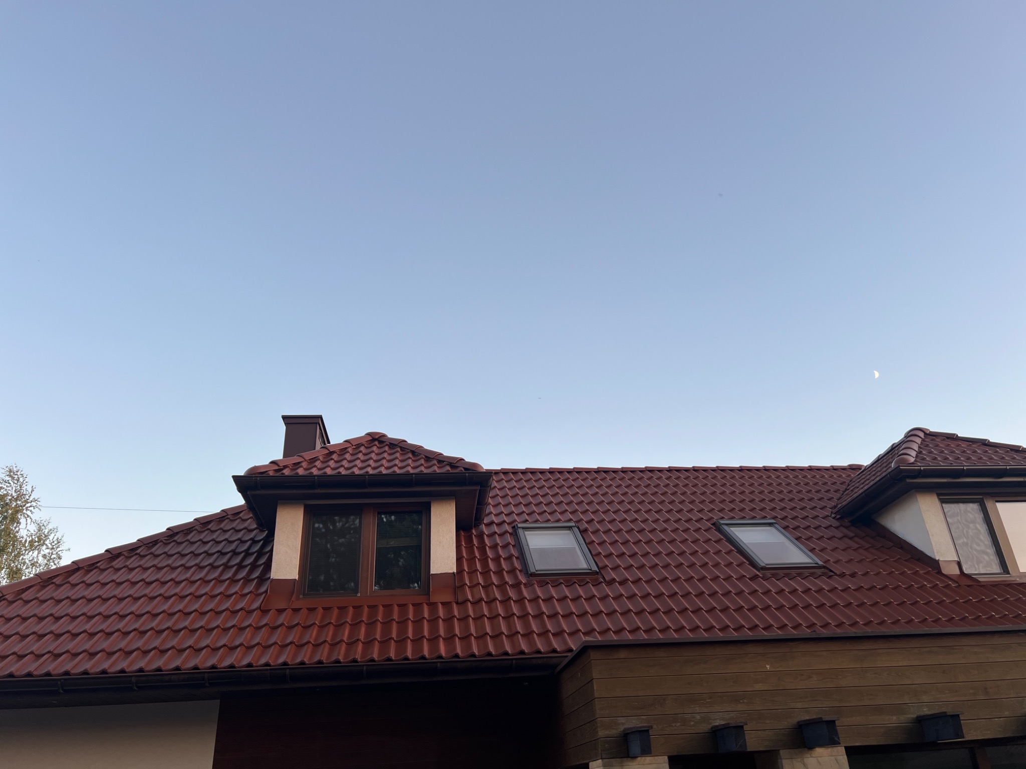 Czerwony dach z dachówką ceramiczną, widoczne okna dachowe i lukarna. Częściowo widoczna elewacja budynku. Błękitne niebo w tle.