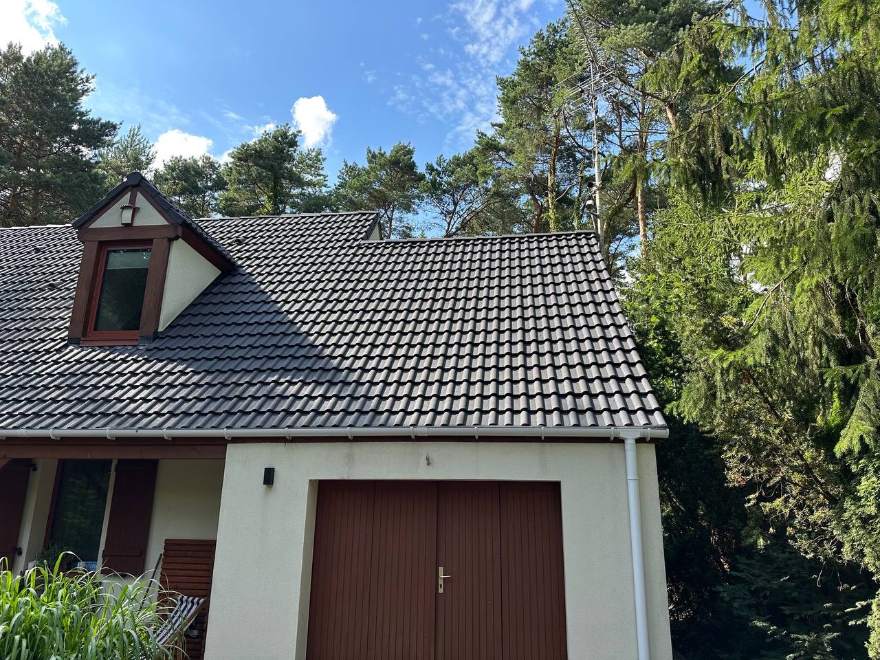Ciemna dachówka ceramiczna na jasnym domu z brązową bramą garażową, otoczonym zielenią drzew i błękitnym niebem z chmurami. Widok z dołu.