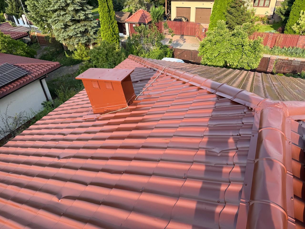 Czerwona dachówka ceramiczna po malowaniu z pomalowaną obudową komina i rynnami. Widok z góry na tle zieleni i zabudowań.