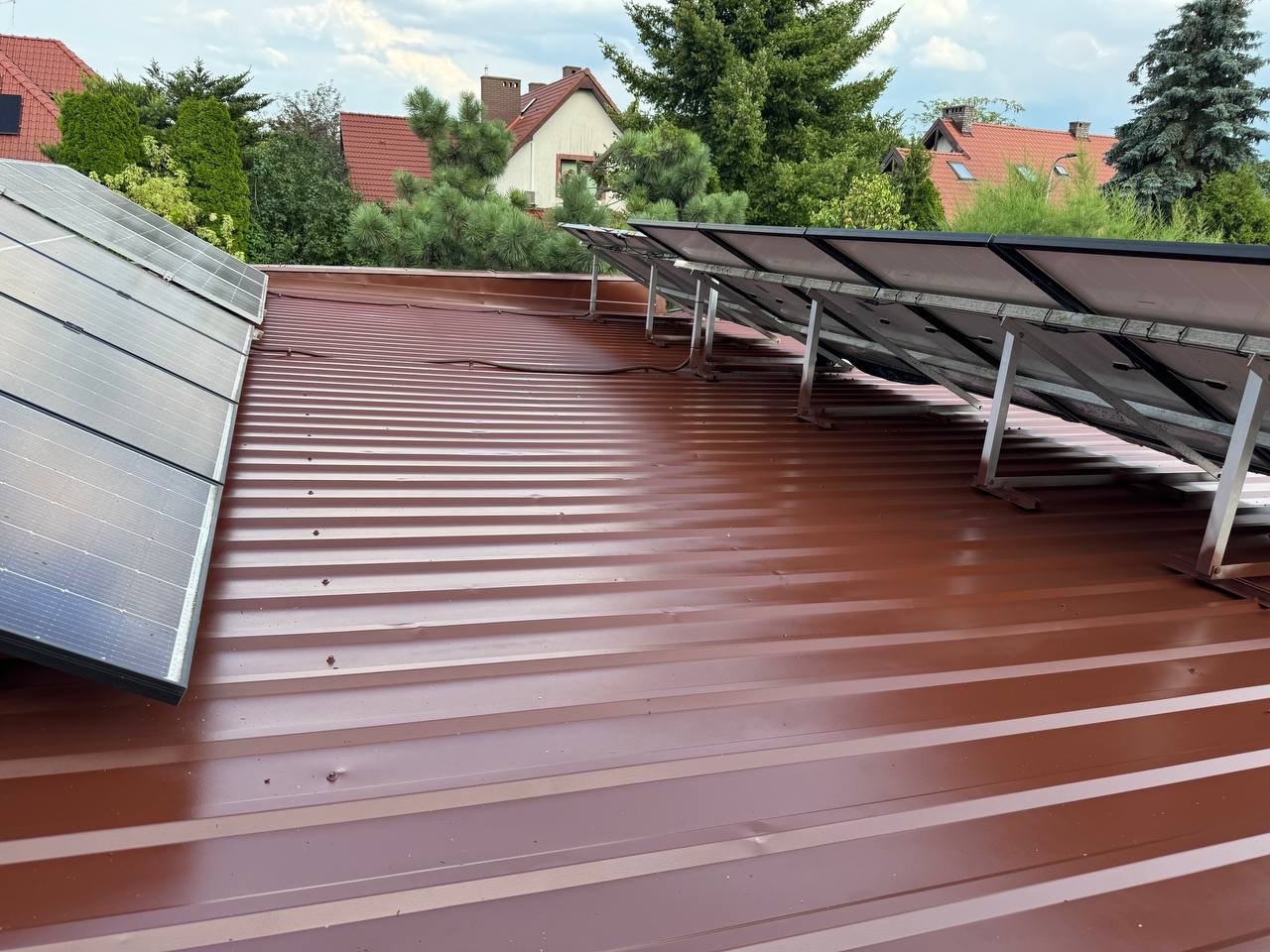 Czerwony dach z blachy trapezowej z zamontowanymi panelami słonecznymi. Widok z góry, na tle zieleni i zabudowań.
